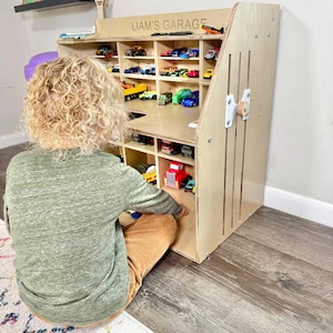 May include: A wooden toy car garage with multiple compartments filled with toy cars of various colors. The garage has the text "LIAM'S GARAGE" on the top. A child is sitting in front of the garage.