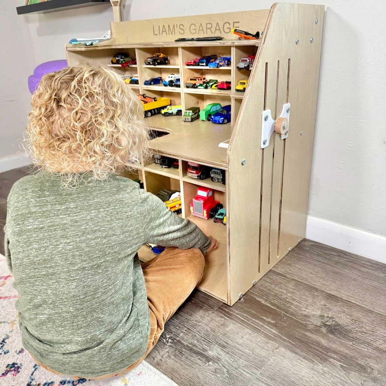 May include: A wooden toy car garage with multiple compartments filled with toy cars of various colors. The garage has the text "LIAM'S GARAGE" on the top. A child is sitting in front of the garage.