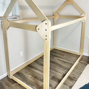 May include: A wooden frame for a bed in the shape of a house. The frame is made of unfinished wood and has a simple design. The frame is sitting on a wooden floor.