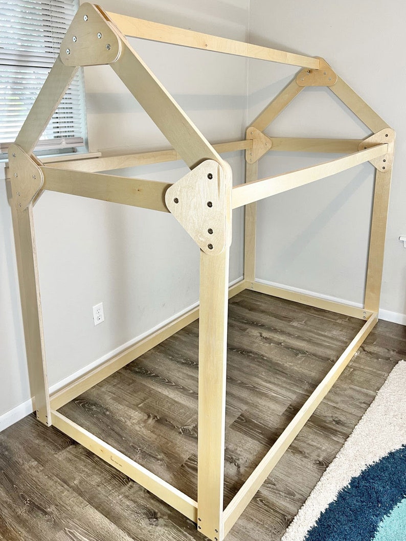 May include: A wooden frame for a bed in the shape of a house. The frame is made of unfinished wood and has a simple design. The frame is sitting on a wooden floor.