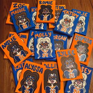 May include: A collection of 12 small canvases painted with portraits of cheerleaders. Each canvas features a different cheerleader's name and a cartoon drawing of them wearing a blue and orange cheerleading uniform. The cheerleaders are all smiling and have their hair pulled back in a ponytail.