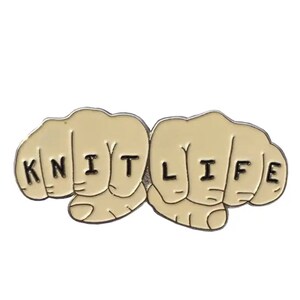 May include: A gold-coloured enamel pin with two fists, each finger spelling out "KNIT LIFE".