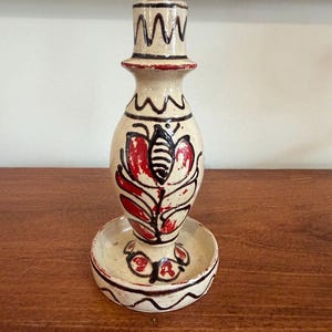 Puede incluir: Un candelero de cerámica color crema con diseños florales rojos y negros. El candelero tiene una base ancha, un cuerpo central en forma de jarrón y un borde decorativo en la parte superior. El diseño está pintado a mano.