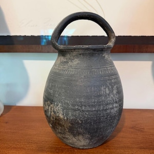May include: A dark gray, weathered ceramic vessel with a rounded body and a single arched handle at the top. The surface features a matte, textured finish with visible signs of wear, including lighter gray patches and horizontal incised lines.