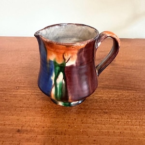 Puede incluir: Una pequeña jarra de cerámica hecha a mano con un cuerpo redondeado y un asa. La jarra presenta una paleta de colores de azul, naranja, verde y granate. El borde es de color gris claro. La jarra está sobre una superficie de madera.