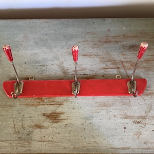 May include: A vintage red wooden coat rack with three metal hooks. Each hook has a red, teardrop-shaped top. The rack is mounted on a weathered, light blue-gray surface.