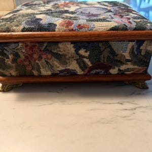 May include: A rectangular wooden box with a floral tapestry design on the sides and top. The box has a dark wood trim and rests on four ornate brass feet. The tapestry features a mix of colors, including blue, red, and green.