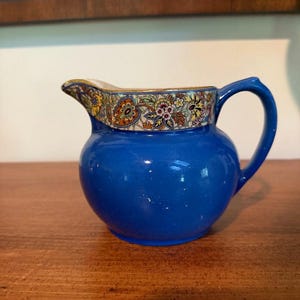 May include: A small, round, cobalt blue ceramic creamer pitcher featuring a decorative band around the top rim. The band displays a colorful, intricate floral and paisley pattern in shades of yellow, red, green, and white with gold trim.
