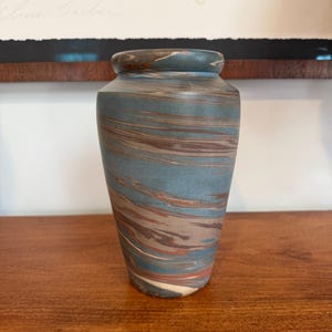 Puede incluir: Un jarrón de cerámica con un diseño veteado azul, marrón y blanco. El jarrón tiene forma cilíndrica con un borde y base ligeramente ensanchados. Los colores se arremolinan, creando un patrón único. El jarrón se exhibe sobre una superficie de madera.