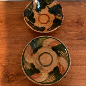May include: Two round ceramic bowls with a tan base color, featuring hand-painted dark green leaf motifs and reddish-brown circular accents in the center. The bowls have a glazed finish with visible variations in color and texture.