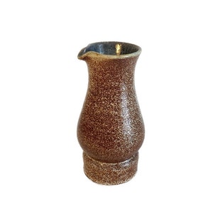 Puede incluir: Una pequeña jarra de cerámica marrón con un esmalte moteado. La jarra tiene un cuerpo redondeado y un pequeño pico.