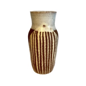 Puede incluir: Un jarrón de cerámica blanco con rayas verticales marrones. El jarrón tiene un fondo redondeado y una abertura ligeramente acampanada.