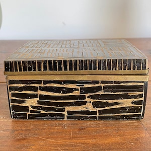 May include: A small, rectangular box made of black wood with a geometric pattern. The box has a brass trim around the lid and a brass hinge.