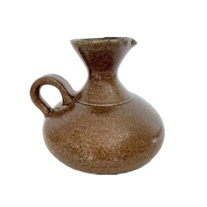 Puede incluir: Una pequeña jarra de cerámica marrón con cuerpo redondeado y asa. La jarra tiene un esmalte moteado y un labio ligeramente acampanado.