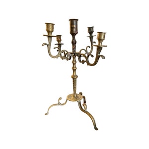 May include: A gold-toned metal candelabra with five candle holders and a three-legged base. The candelabra has a decorative, ornate design.