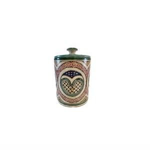 May include: A white ceramic jar with a lid, decorated with green, blue, pink, and yellow geometric patterns. The jar has a heart-shaped design in the centre.
