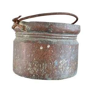 May include: A large, copper pot with a worn, aged patina. The pot has a single handle with a hinged latch. The pot is likely used for cooking or decorative purposes.