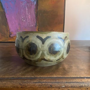 May include: A ceramic bowl with a rounded shape and a textured surface. The bowl is primarily olive green with a repeating pattern of dark brown circles and stylized black shapes. The bowl sits on a wooden surface.