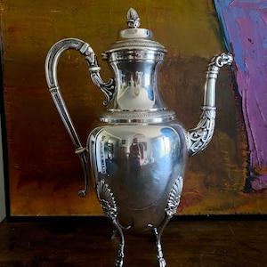 May include: A highly polished silver coffee pot with intricate detailing. It has a curved handle, a decorative spout, and stands on three small feet. The lid is topped with a finial. The pot reflects the surrounding environment.
