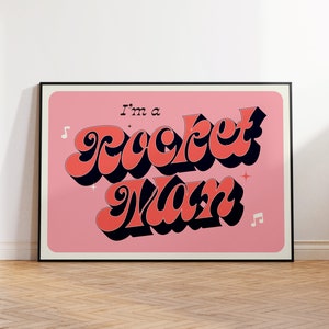 May include: A framed poster featuring the text "I'm a Rocket Man" in a retro style. The coral and black text is set against a pink background, with musical notes and stars. The poster is in a black frame.