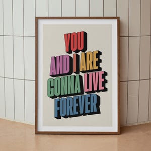 May include: A framed art print featuring the phrase "YOU AND I ARE GONNA LIVE FOREVER" in vibrant, 3D block letters. The text is set against a cream background, within a brown wooden frame.