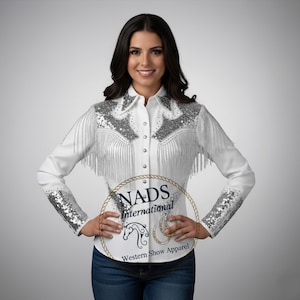 Puede incluir: Camisa blanca de estilo western con detalles de lentejuelas plateadas en los hombros y puños, y flecos. La camisa tiene una botonadura frontal y un cuello. La imagen incluye el texto "NADS International Western Show Apparel".