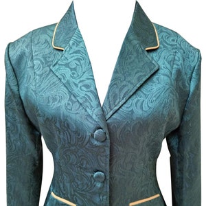 Puede incluir: Una chaqueta de color verde azulado con estampado paisley y ribete crema. La chaqueta tiene un cierre de botón único y dos bolsillos. El texto "NADS INTERNATIONAL" está impreso en la parte inferior de la chaqueta.