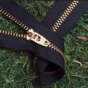 May include: A black zipper with metallic gold-toned teeth and a matching gold-toned metal slider pull. The slider is clearly embossed with the letters YKK. The zipper tape is made of a dark black fabric, and the teeth are arranged in two rows.