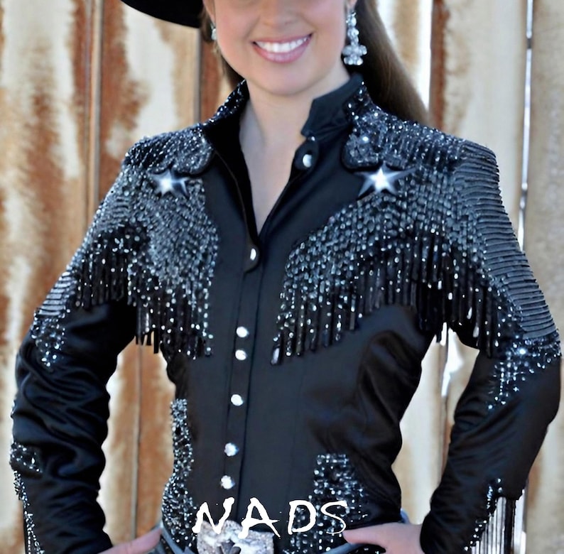 May include: A black satin shirt with a beaded design and fringe. The shirt has a button-up front and long sleeves. The shirt is decorated with rhinestones and the word "NADS" is visible on the front.