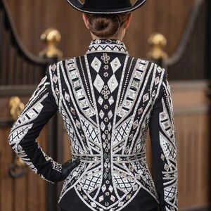 May include: A black long-sleeved show jacket featuring intricate white and silver geometric patterns, embellished with numerous clear crystals and rhinestones in diamond and circular shapes, paired with a matching black cowboy hat with silver trim.