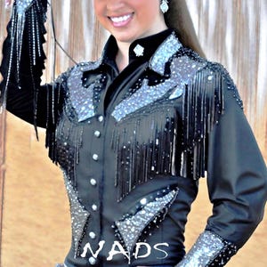 May include: Black western-style shirt with silver embellishments and fringe. The shirt has a button-up front and long sleeves. The word "NADS" is visible on the bottom of the shirt.