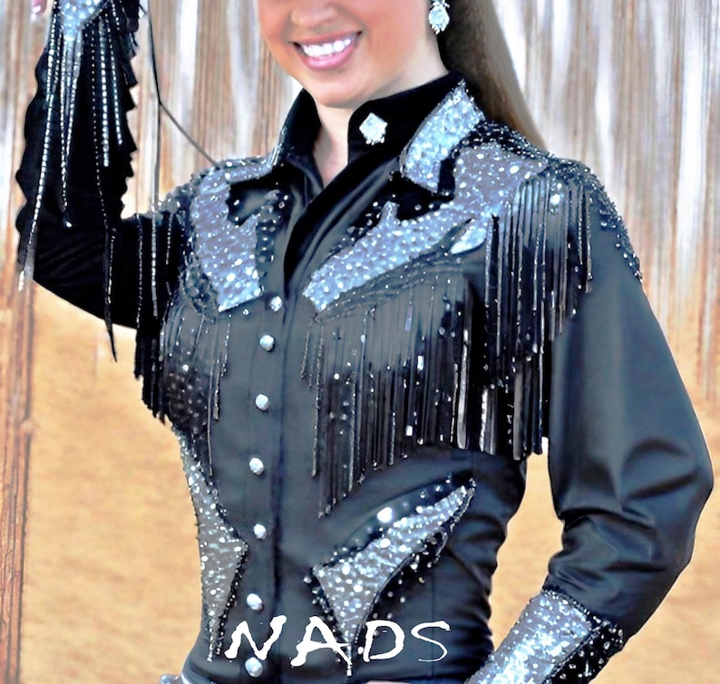 May include: Black western-style shirt with silver embellishments and fringe. The shirt has a button-up front and long sleeves. The word "NADS" is visible on the bottom of the shirt.