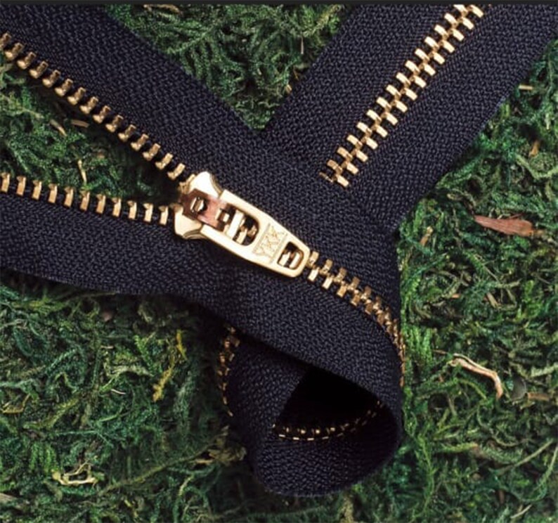 May include: A black zipper with a gold metal slider and teeth. The zipper is lying on a bed of green moss.
