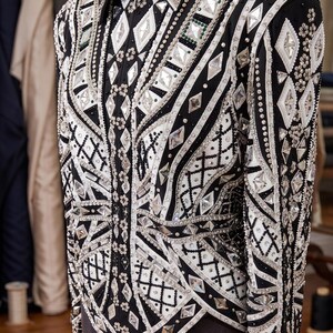 May include: A black long-sleeve jacket featuring an intricate pattern of white fabric panels, silver-toned metallic accents, and numerous clear, diamond-shaped crystals arranged in geometric designs across the front and sleeves.