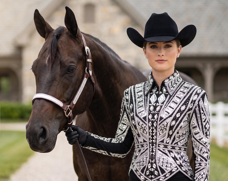 May include: A dark brown horse with a leather halter and a person wearing a black cowboy hat and a long-sleeved black shirt heavily embellished with intricate silver and clear crystal geometric patterns across the front and sleeves.
