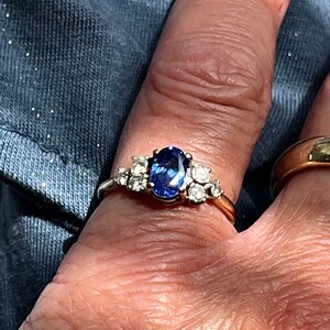 Op de afbeelding: Een gouden ring met een blauwe ovale edelsteen omringd door kleinere heldere edelstenen.