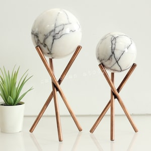 Puede incluir: Dos esferas de mármol blanco con vetas grises, cada una sentada en un soporte de trípode de color cobre.