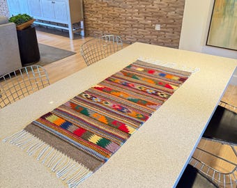 May include: A rectangular, colourful woven table runner with a geometric pattern. The runner has a brown border and white fringe on both ends. The design includes stripes of red, orange, green, blue, and purple. It is placed on a light-coloured table.