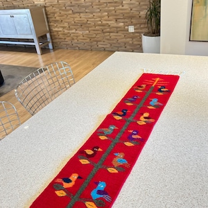 Peut inclure: Un chemin de table rouge avec un motif tissé d'un arbre avec des oiseaux colorés. L'arbre est vert et les oiseaux sont orange, bleu, violet et jaune. Le chemin de table a des franges blanches aux extrémités.
