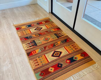 May include: A rectangular, woven rug with a tan border and fringed edges. The rug features a repeating pattern of geometric shapes in red, orange, blue, green, and brown. The rug is placed on a light wood floor.