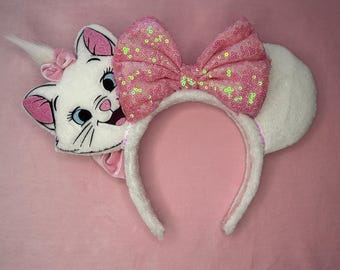 May include: White plush headband featuring a cartoon cat face with pink accents and a large pink sequined bow. The cat has blue eyes and a small pink bow. The ears are white and fluffy, and the headband is pink.