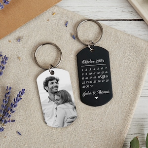 Può includere: Due portachiavi personalizzati. Uno presenta una foto in bianco e nero di una coppia. L'altro è nero con il testo "Oktober 2024" e un calendario che evidenzia una data, con i nomi "Julia & Thomas".