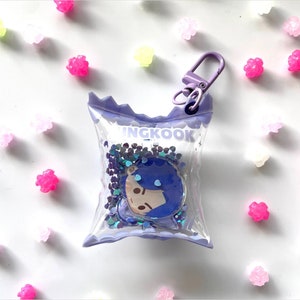 May include: A clear plastic keychain shaped like a purple snack bag with a cartoon character inside. The bag has the word "INGKOOK" printed on it and is filled with iridescent glitter.