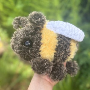 May include: A plush bee toy crafted from fuzzy yarn with alternating horizontal stripes of dark brown and bright yellow. The bee features two smooth black plastic eyes and a pair of textured, white crocheted wings attached to its back.