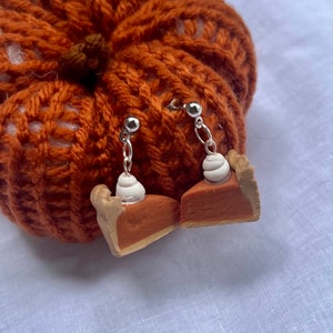 May include: A pair of pumpkin pie slice earrings with white whipped cream topping. The earrings are made of polymer clay and have silver hooks.