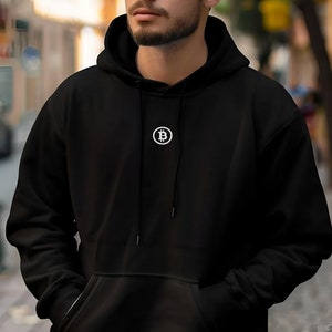 Puede incluir: Una sudadera con capucha negra con un logotipo de Bitcoin blanco en la parte delantera.