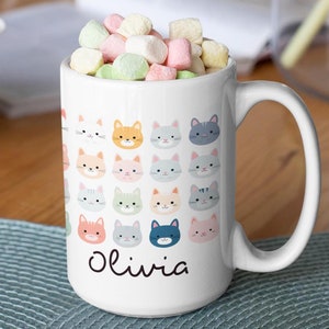 May include: White ceramic mug filled with colorful marshmallows. The mug is decorated with a pattern of various cat faces in pastel colors. The name "Olivia" is written in black script on the mug.