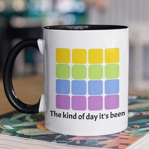 Peut inclure: Une tasse en céramique blanche avec un bord et une poignée noirs. La tasse présente un motif coloré d'une grille de carrés en jaune, vert, bleu et violet. Le texte "The kind of day it's been" est imprimé sous le motif.