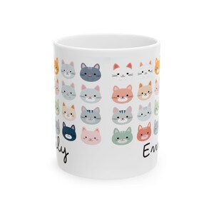 May include: White ceramic mug decorated with a repeating pattern of colorful cat faces. The cat faces are in various colors, including orange, gray, pink, and blue. The names "Emily" and "Lily" are written on the mug in black script.