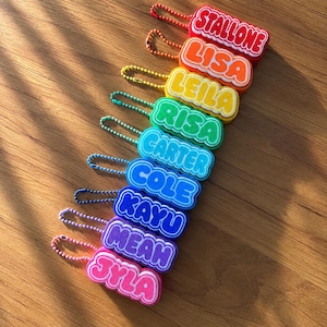 May include: A collection of ten colorful silicone name keychains arranged in a diagonal line. Each keychain features a unique name in white bubble letters on a vibrant background, including Stallone, Lisa, Leila, Risa, Carter, Cole, Kayu, Meah, and Jyla.